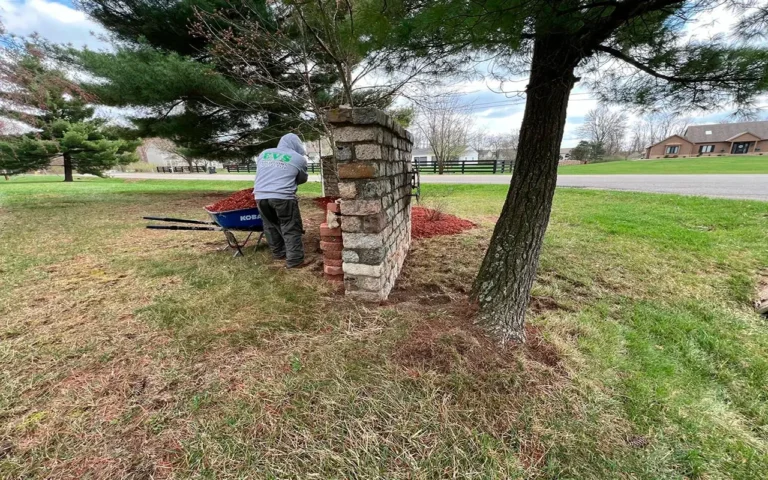 A worker wearing an "EVS Landscaping" hoodie stands next to a blue wheelbarrow filled with red mulch, preparing bricks near a stone structure as part of a Spring CleanUp in Ohio.
