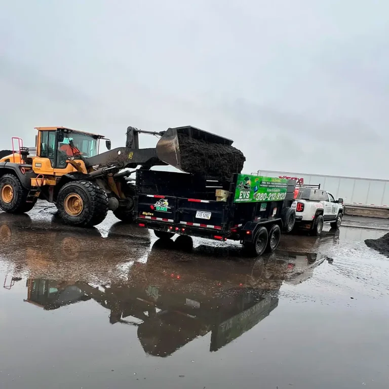A yellow front loader dumps a load of soil into a black trailer attached to a white pickup truck, preparing materials for a Spring CleanUp in Ohio. The wet ground reflects the vehicles and equipment, showcasing an active work site.