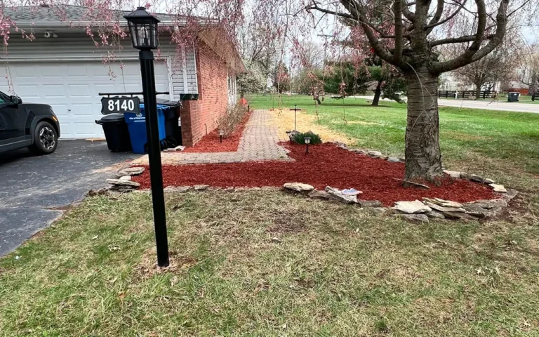 A suburban front yard featuring a landscaped area with red mulch, rocks, and a tree, along with a driveway, a brick house, and a black car, captured as part of a Spring CleanUp in Ohio.
